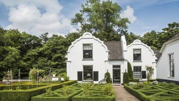 A white building with a symmetrical facade, a central entrance, and a well-manicured garden with hedges and a brick pathway.