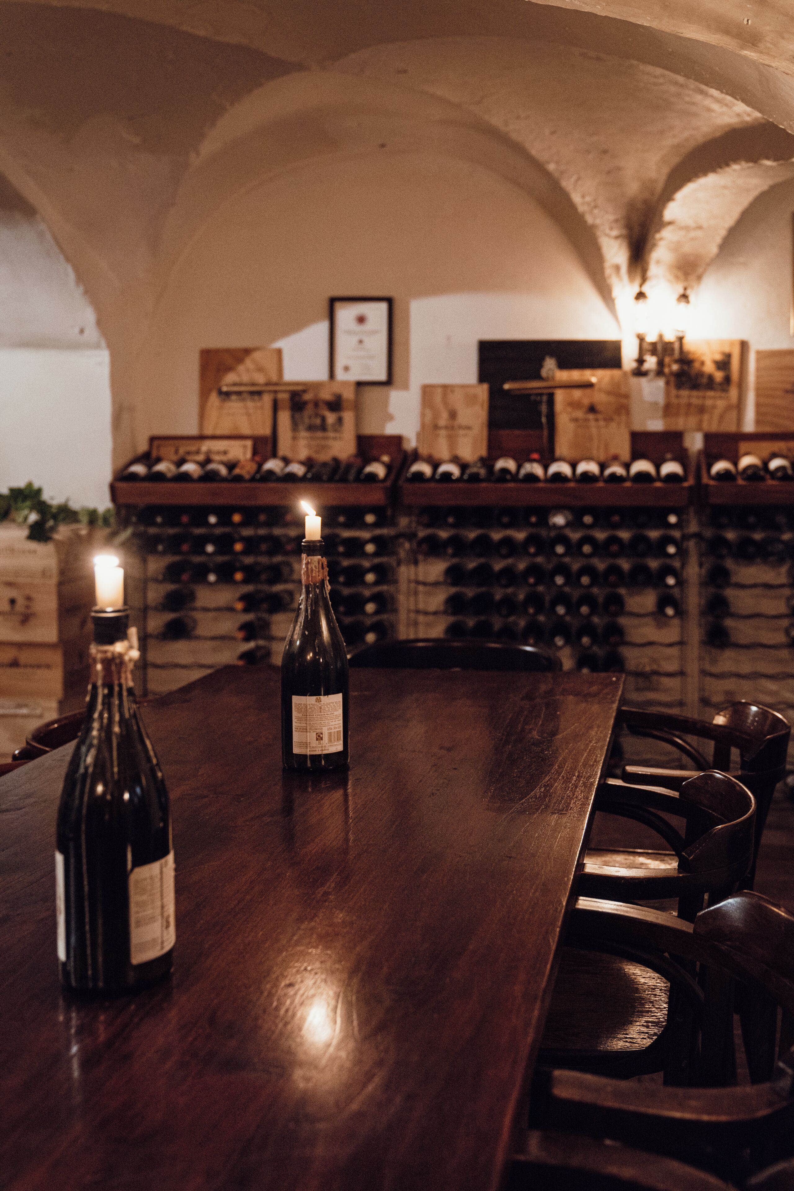 A wooden table with two bottles of wine and a lit candle, set in a dimly lit room with arched ceilings and a wine rack in the background.