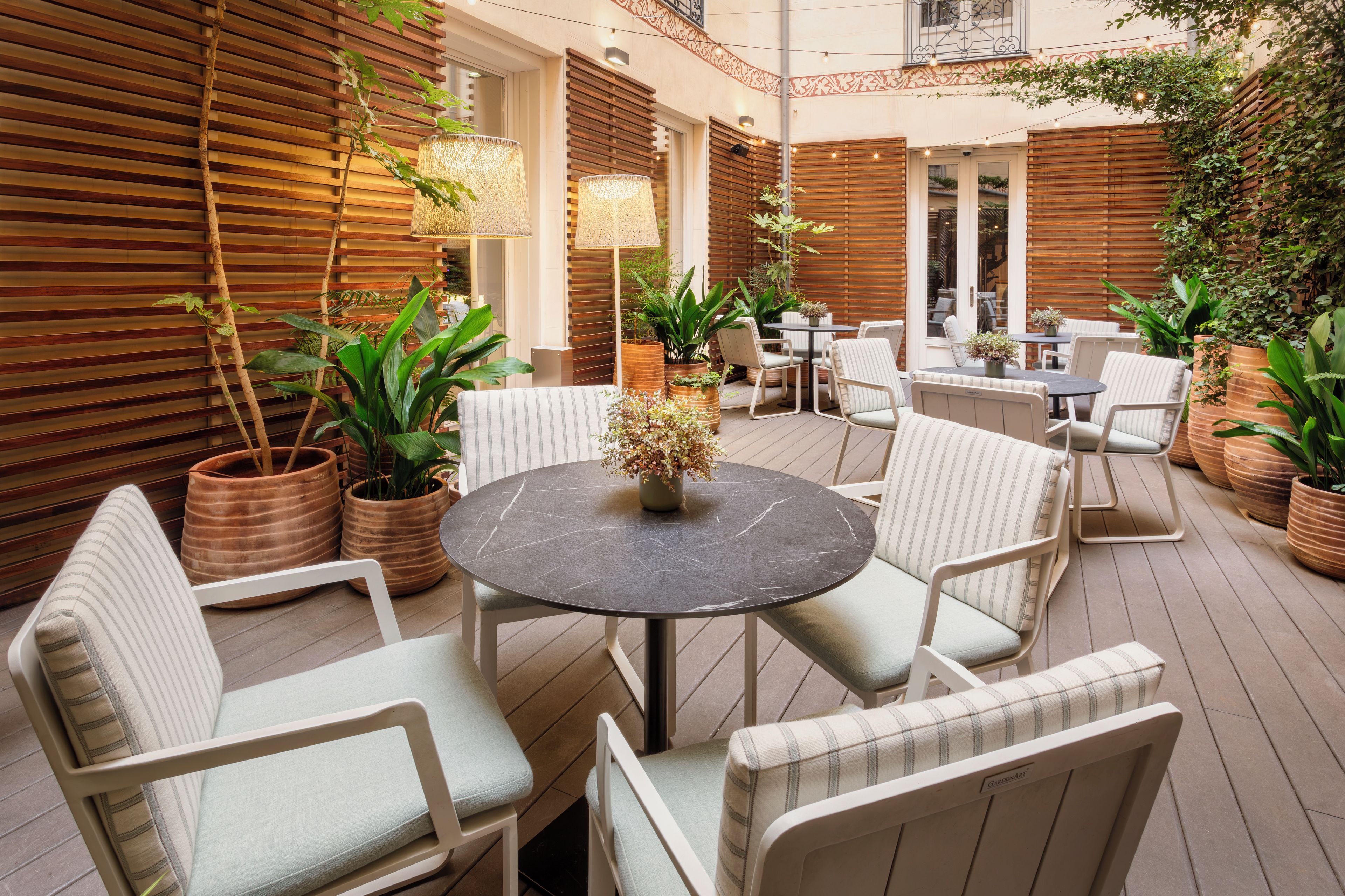 Un patio con una mesa redonda rodeada de sillas, plantas en macetas y paredes de listones de madera.