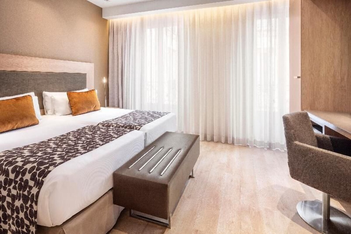 A hotel room with a bed, a bench, a chair, and a large window with curtains.