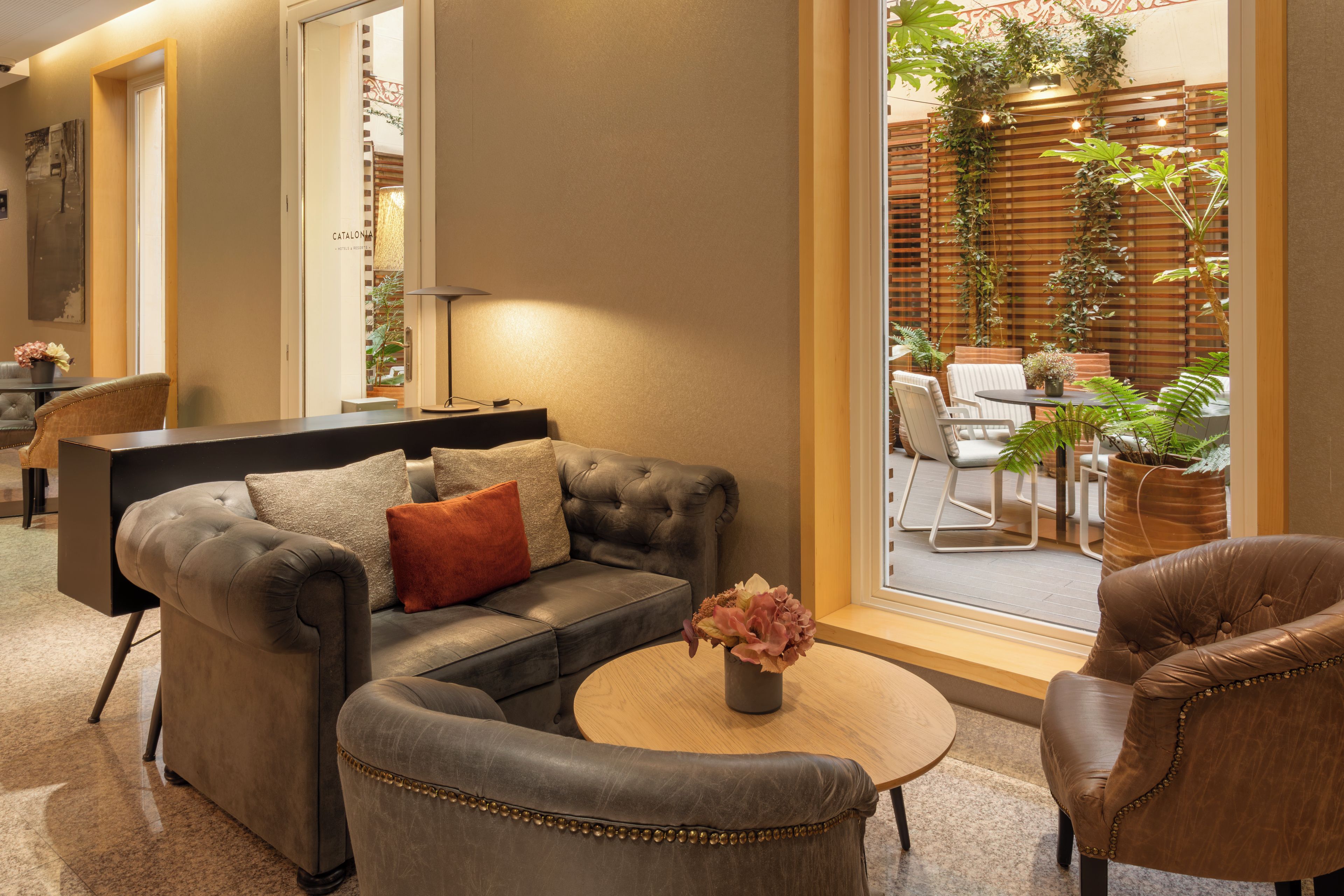 A modern living room with a sofa, armchair, and coffee table, featuring a view of an outdoor patio with plants and seating.