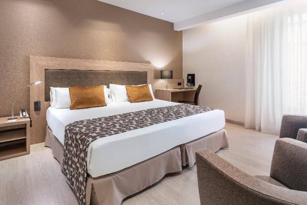 A hotel room with a large bed, a desk, a chair, and a bedside table.