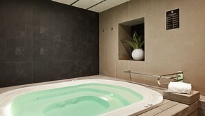 A modern spa room with a large freestanding bathtub, a small potted plant, and a sign indicating amenities. - Catalonia Plaza Mayor (Madrid)