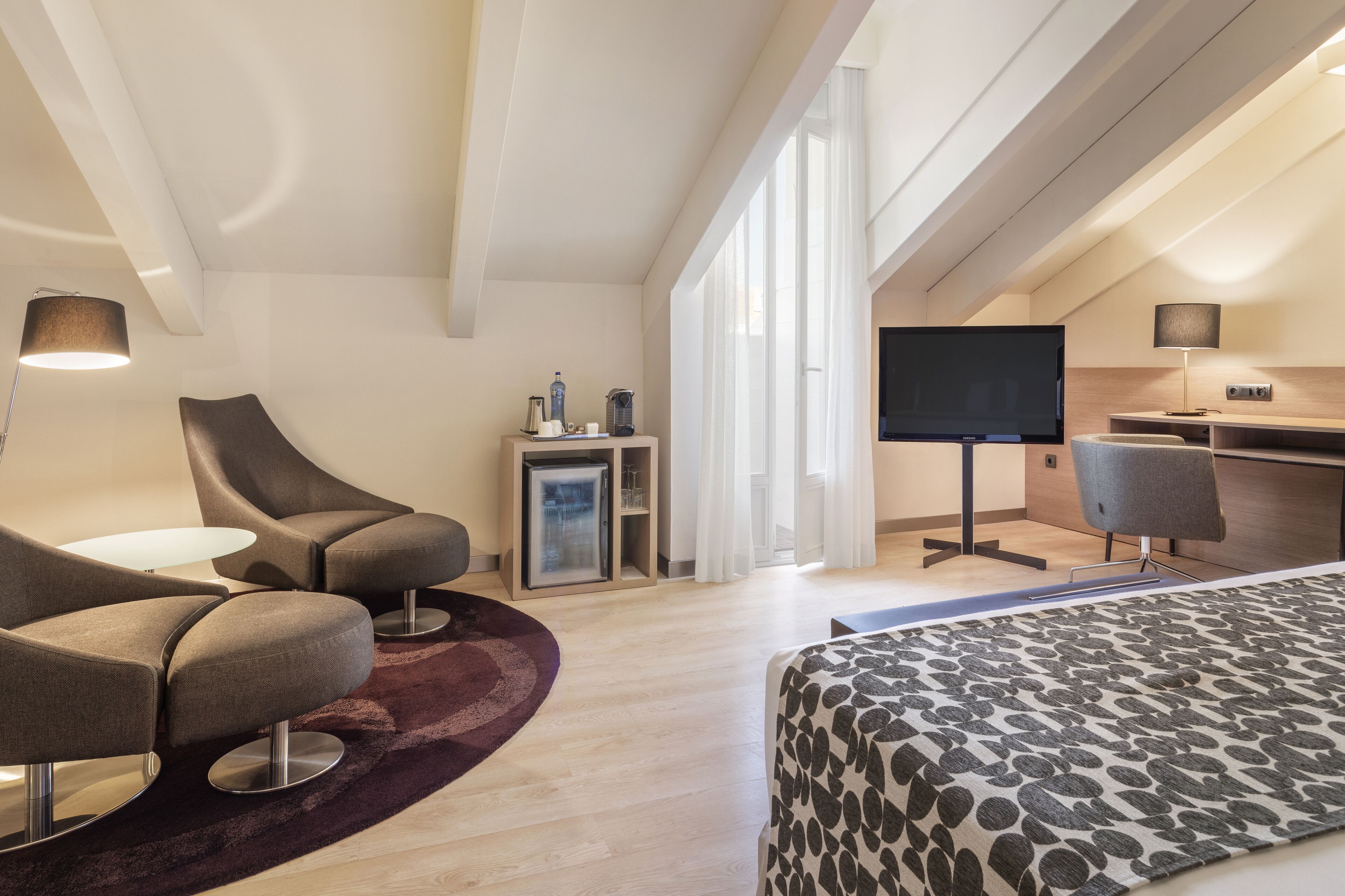 Moderni hotellihuone, jossa on kaareva sohvaryhmä, kuviollinen päiväpeite, pieni keittokomero, litteänäyttöinen televisio ja suuret ikkunat.