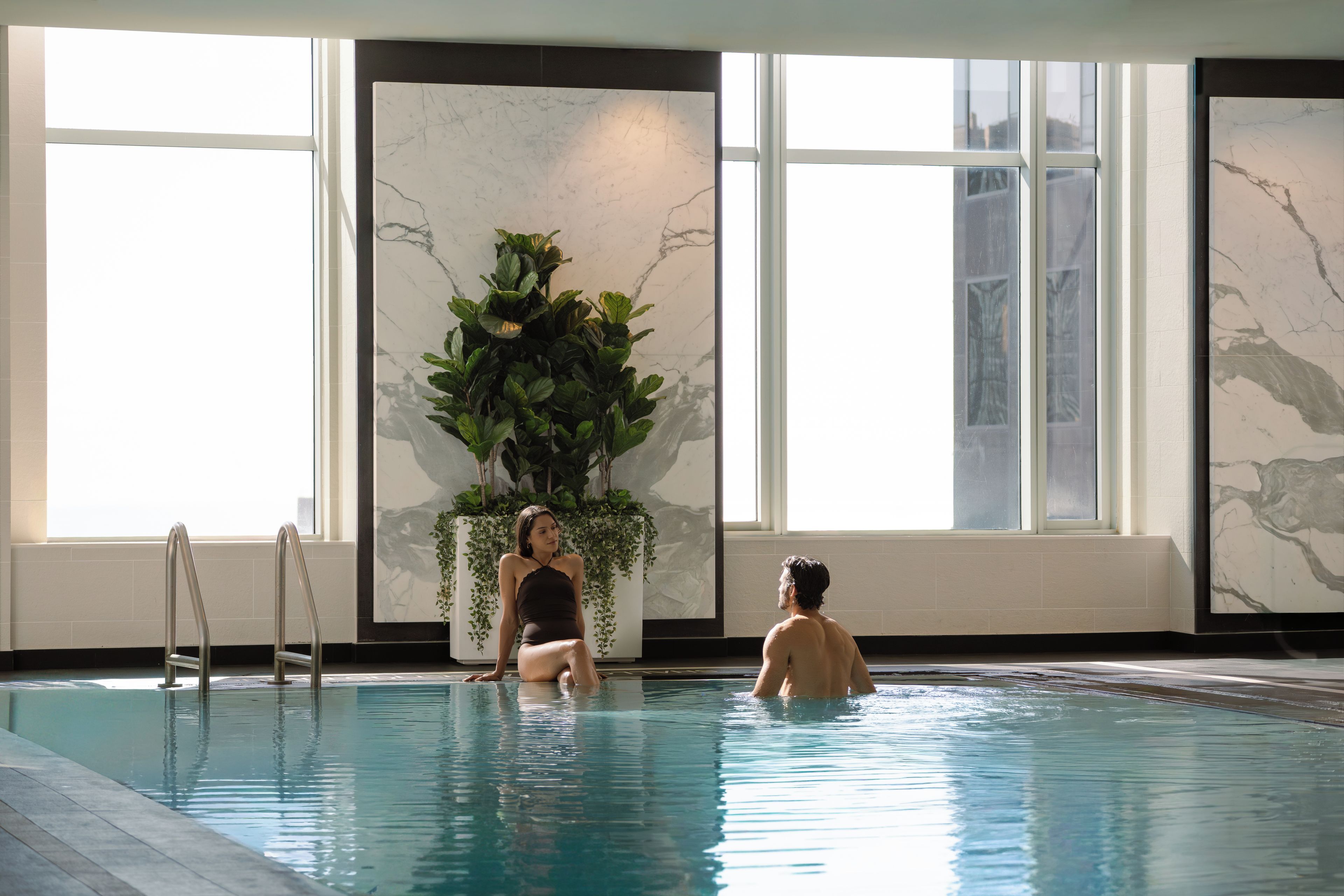 An indoor swimming pool with clear water, a plant decoration, and a view of the outside through large windows.