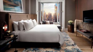 A modern hotel room with a large bed, a TV, a desk with a lamp, and a view of the cityscape through a large window.