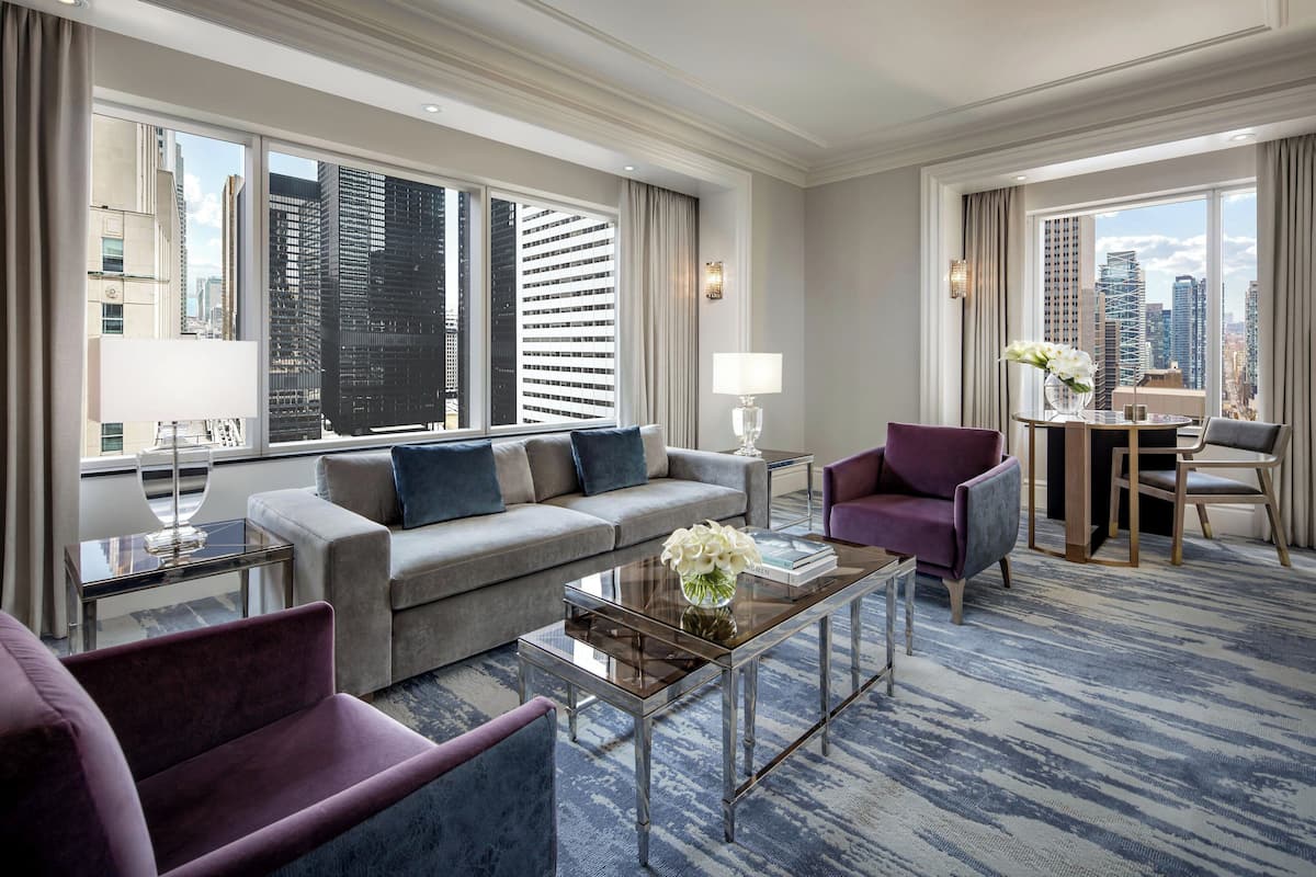 Une chambre d’hôtel moderne avec un canapé gris, un fauteuil violet et une table basse en verre. De grandes fenêtres offrent une vue sur la ville. On y trouve également un coin repas avec une table et des chaises, ainsi qu’une lampe sur une table d’appoint.