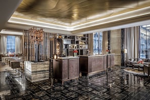 A modern hotel lobby with a marble reception desk, dark wood furniture, and a large window offering a city view.