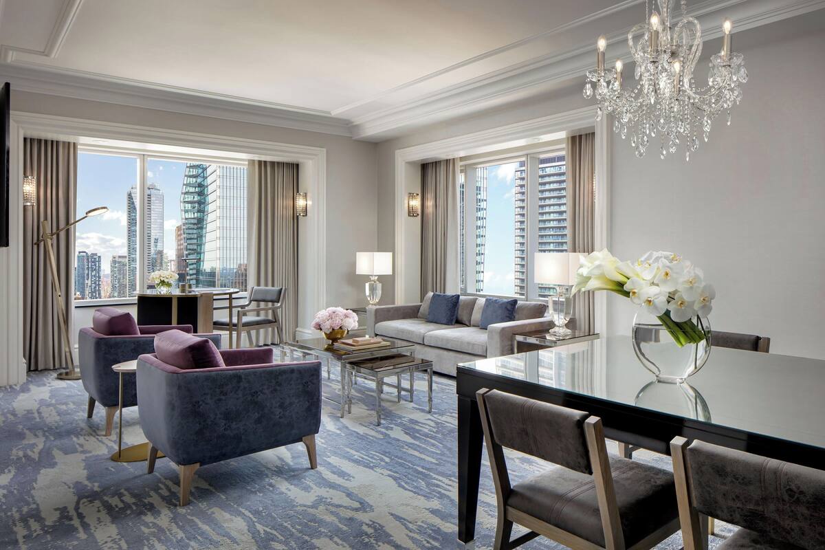 Une chambre d’hôtel moderne avec vue sur la ville, un coin repas avec une table en verre et un lustre.