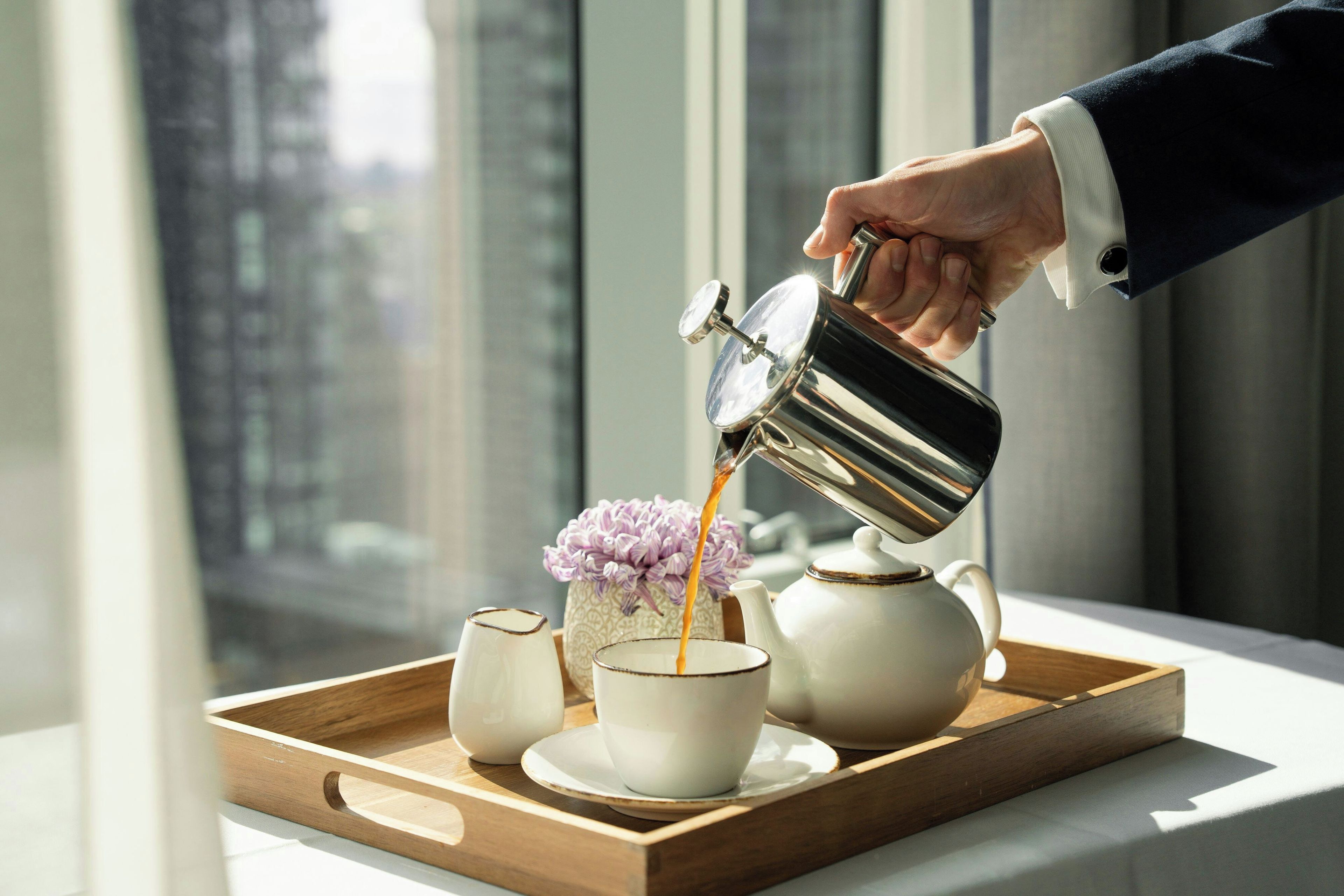 Una mano vertiendo café en una taza sobre una bandeja con una tetera y un florero.