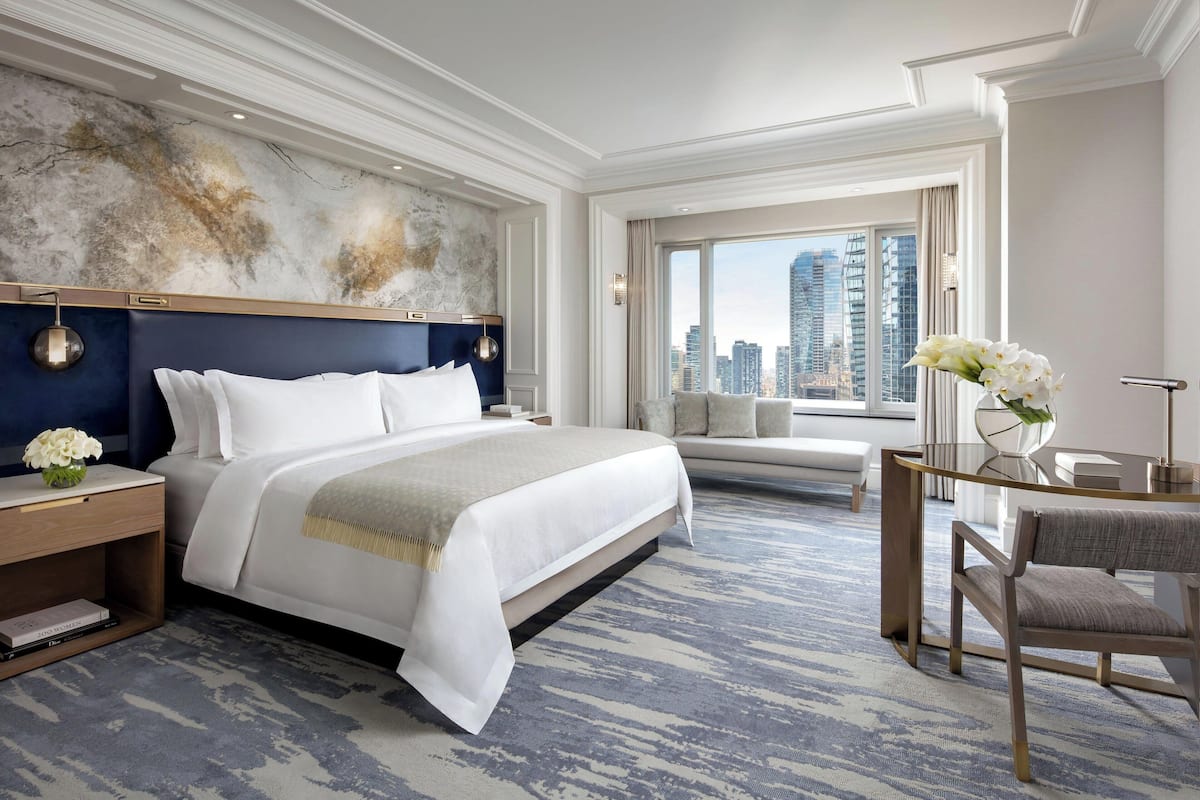 Une chambre d’hôtel moderne dotée d’un grand lit, d’une vue sur la ville, d’un coin salon et d’une petite table sur laquelle est posé un vase de fleurs.