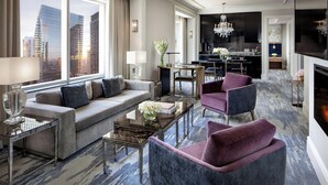 A modern hotel room with a large window offering a city view, a living area with a sofa, armchairs, and a coffee table, and a separate dining area with a chandelier.