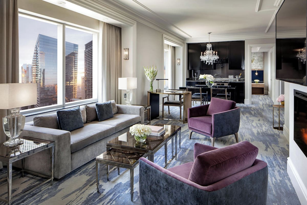 Une chambre d’hôtel moderne dotée d’une grande fenêtre donnant sur la ville, d’un coin salon avec un canapé, des fauteuils et une table basse, ainsi que d’un espace repas séparé agrémenté d’un lustre.