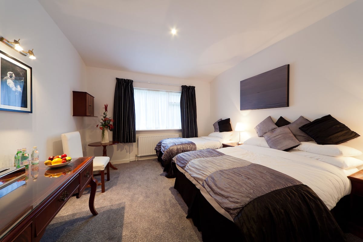 A hotel room with two beds, a desk with a chair, a small table with a vase, and a window with curtains.