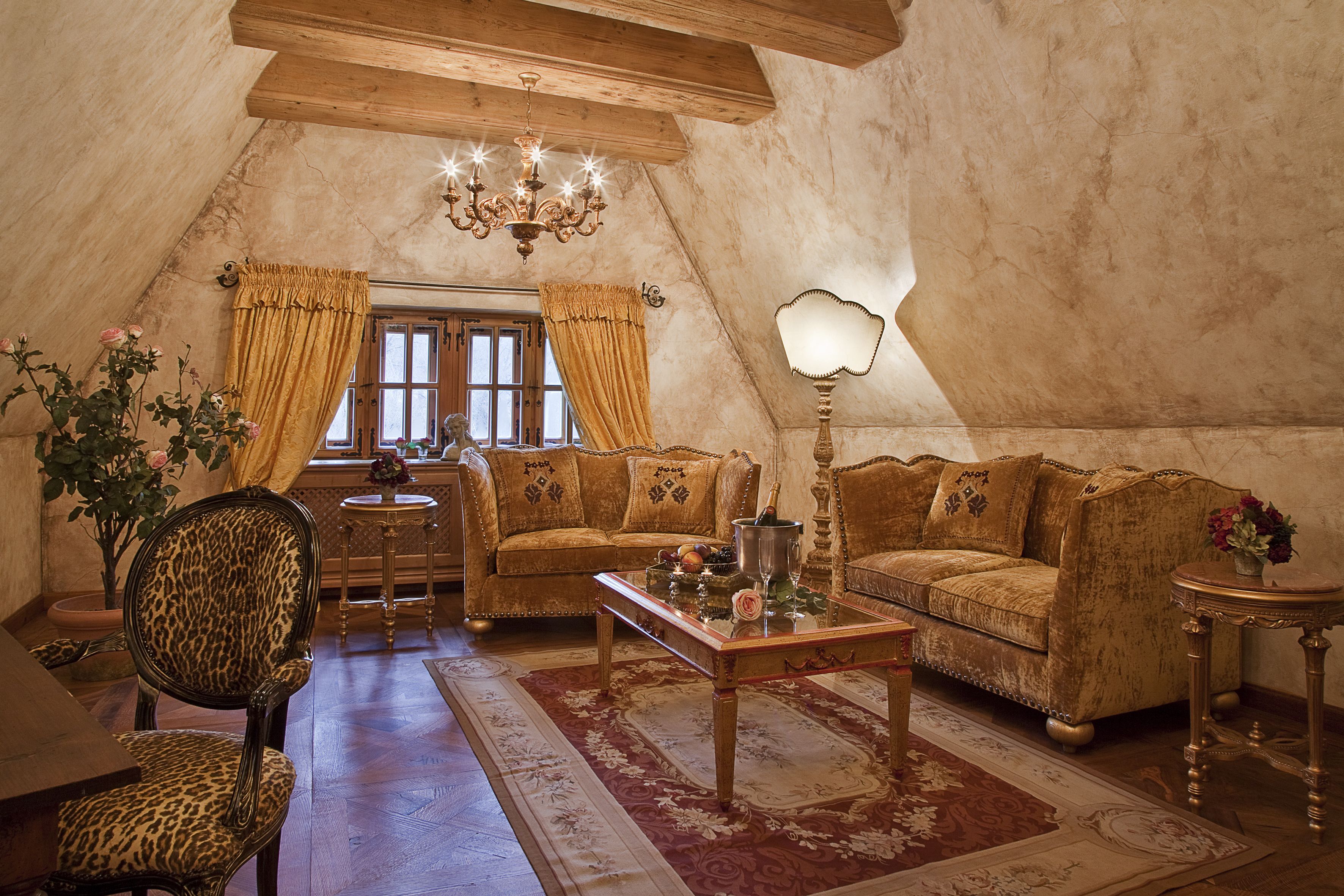 A cozy attic room with a wooden ceiling, a chandelier, a sofa set, a leopard print chair, a coffee table, and a lamp.