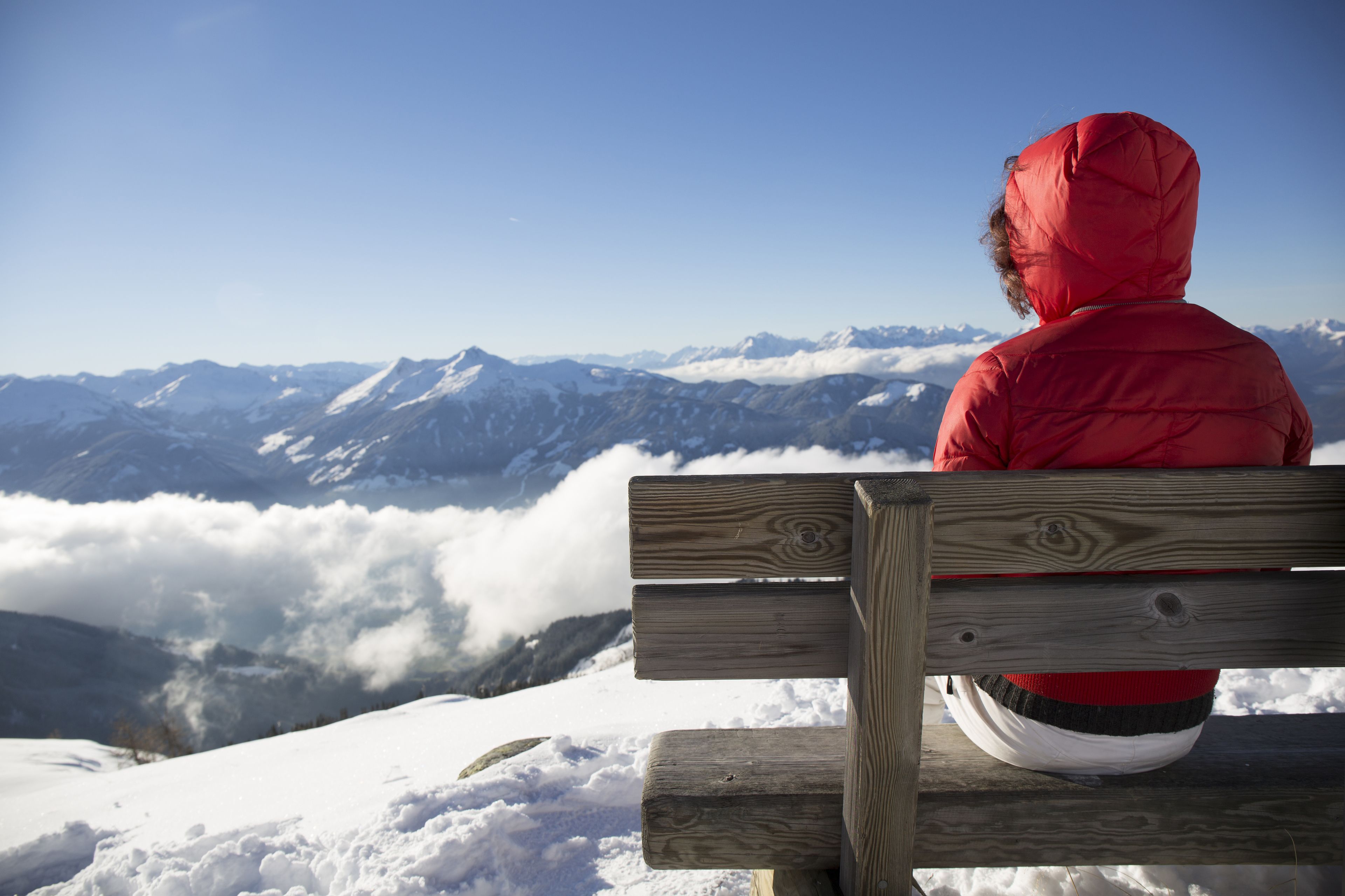 Una persona con una chaqueta roja sentada en un banco de madera contemplando un paisaje montañoso nevado.