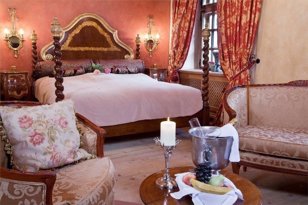 Un dormitorio con cama de dos columnas, sillones de estampado floral, una mesita de noche con una vela y un frutero.