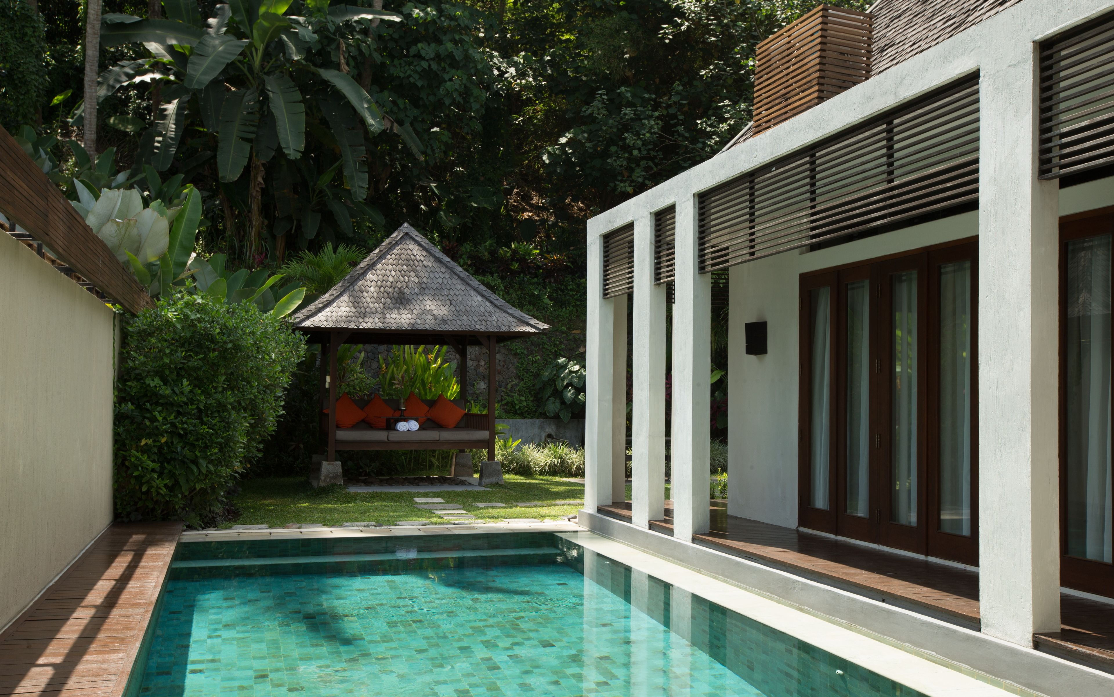 A modern pool area with a small gazebo, surrounded by lush greenery and a white building with large windows.