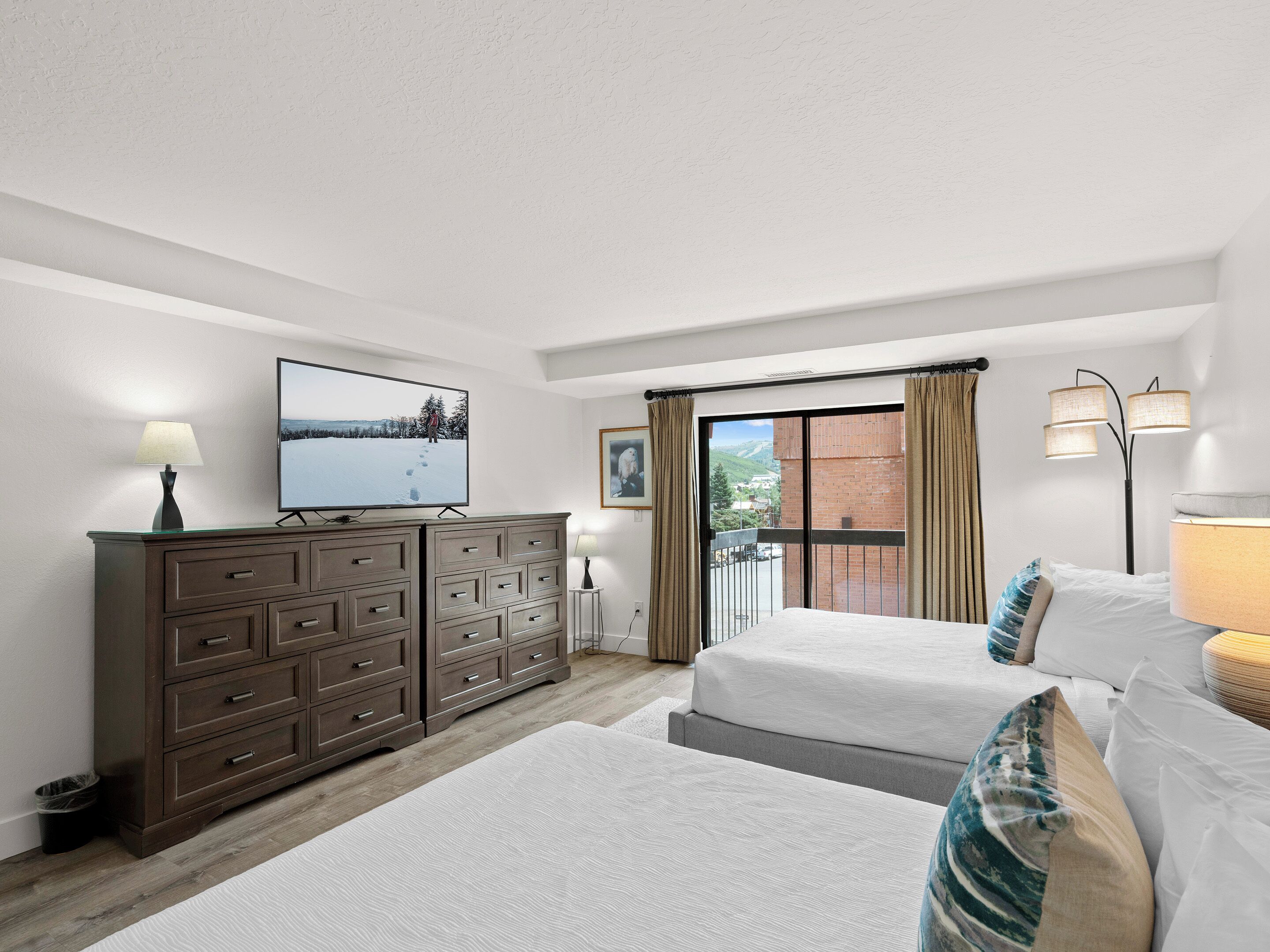 Een slaapkamer met twee bedden, een dressoir, een televisie en een balkon met gordijnen.