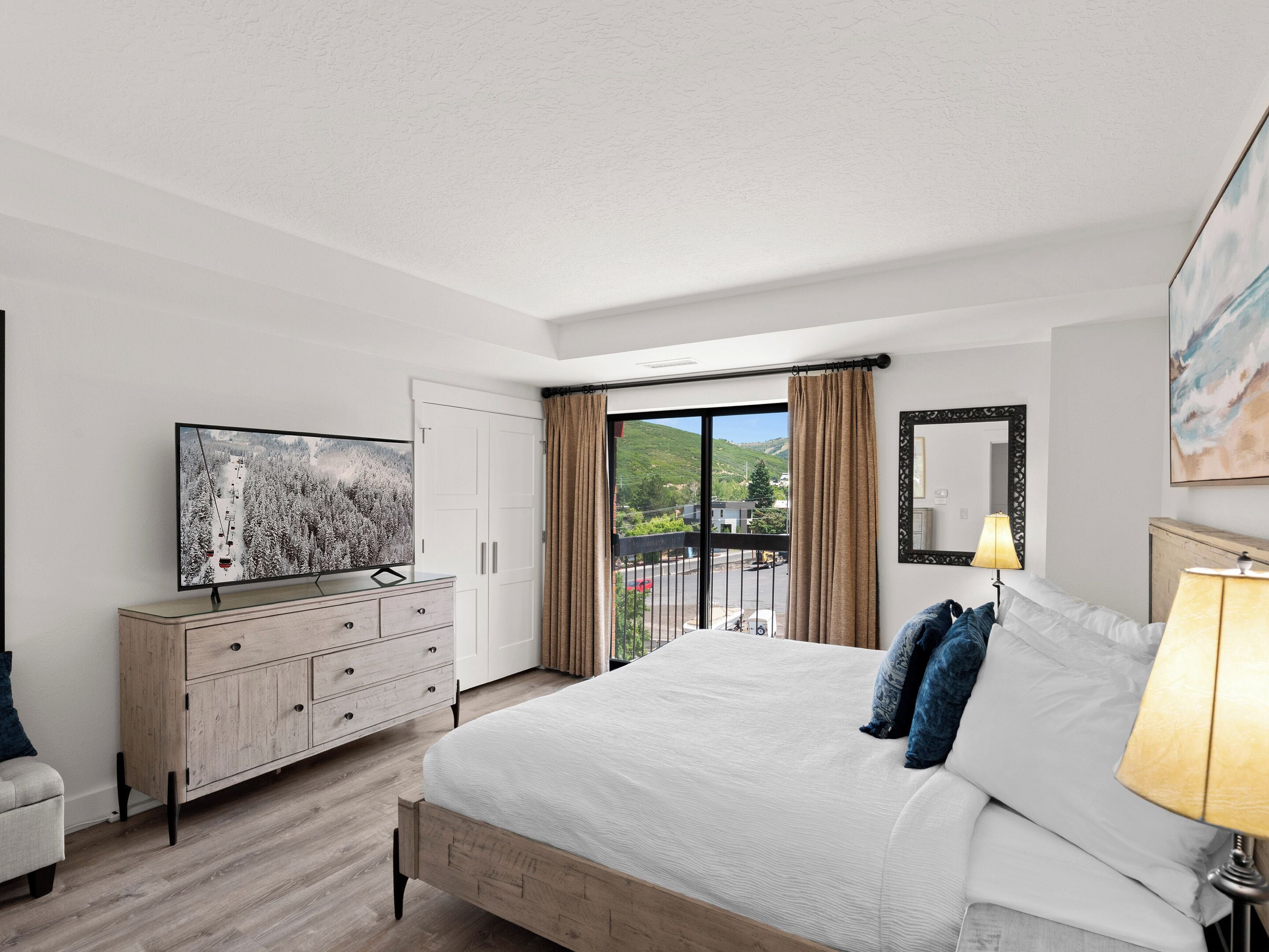 A hotel room with a bed, a TV, a dresser, a balcony with a view of the outside, and a painting on the wall.