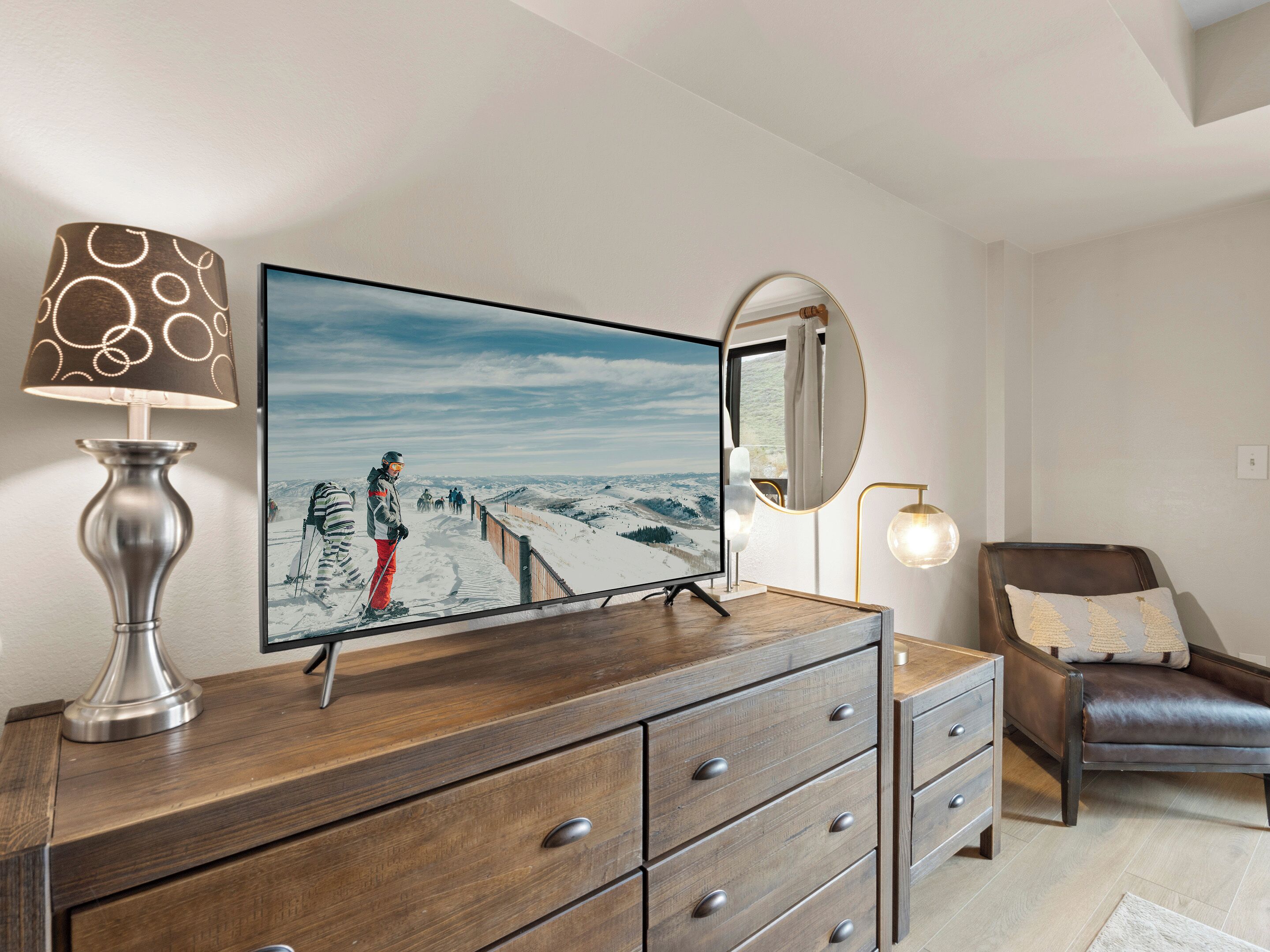 A room with a wooden dresser, a television displaying a snowy landscape, a mirror, and a chair.