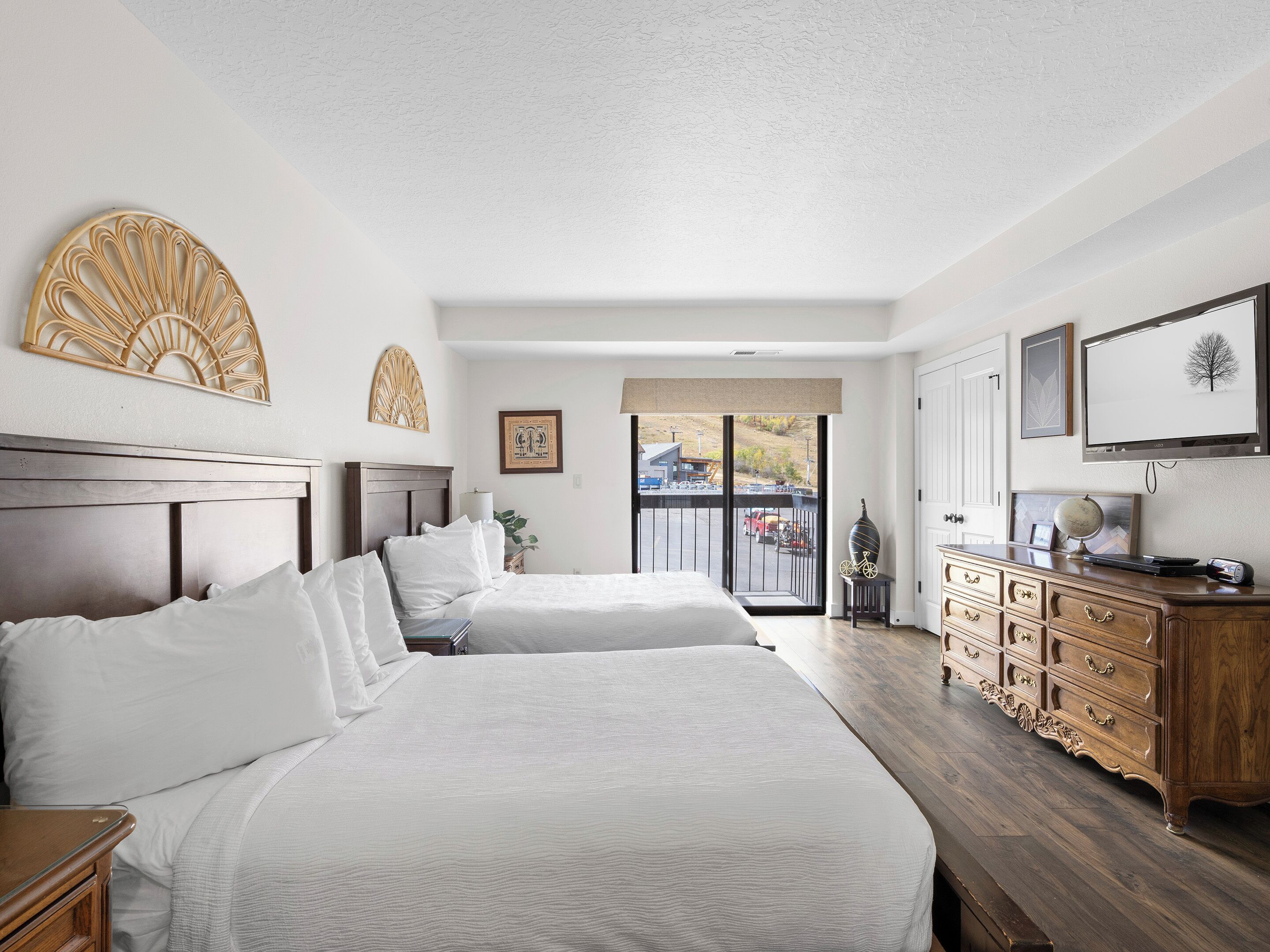 Quarto de hotel com duas camas, cómoda de madeira, televisão e varanda com vista para o exterior.