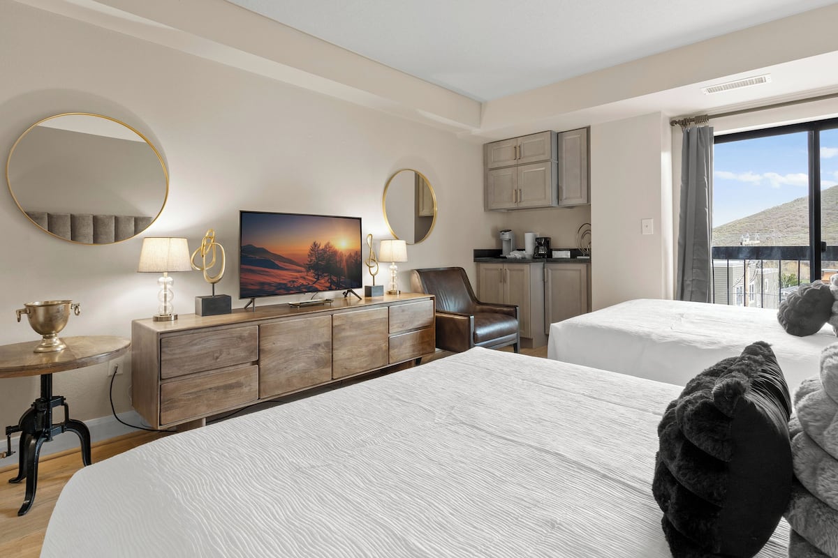 Habitación de hotel con dos camas, televisor, escritorio con lámpara, silla, cómoda y vista al exterior.