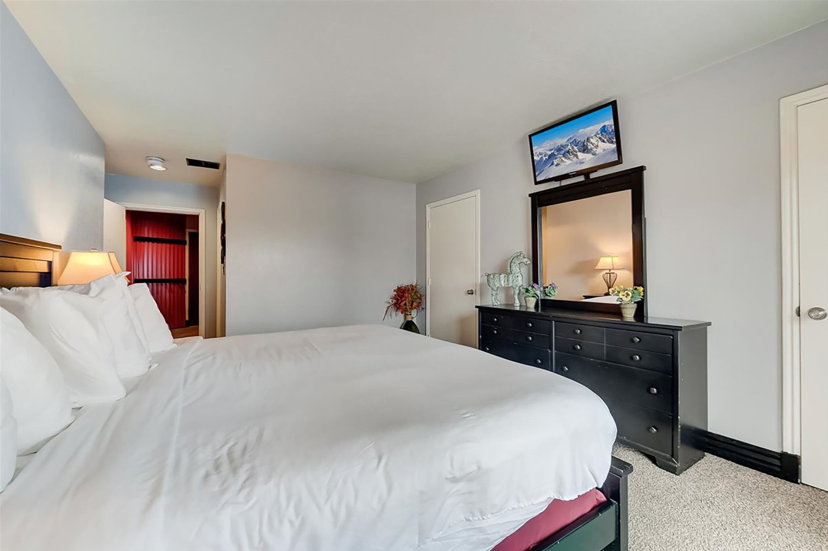 Una habitación de hotel con una cama, un aparador, un televisor montado sobre el aparador y una puerta que da a otra habitación.