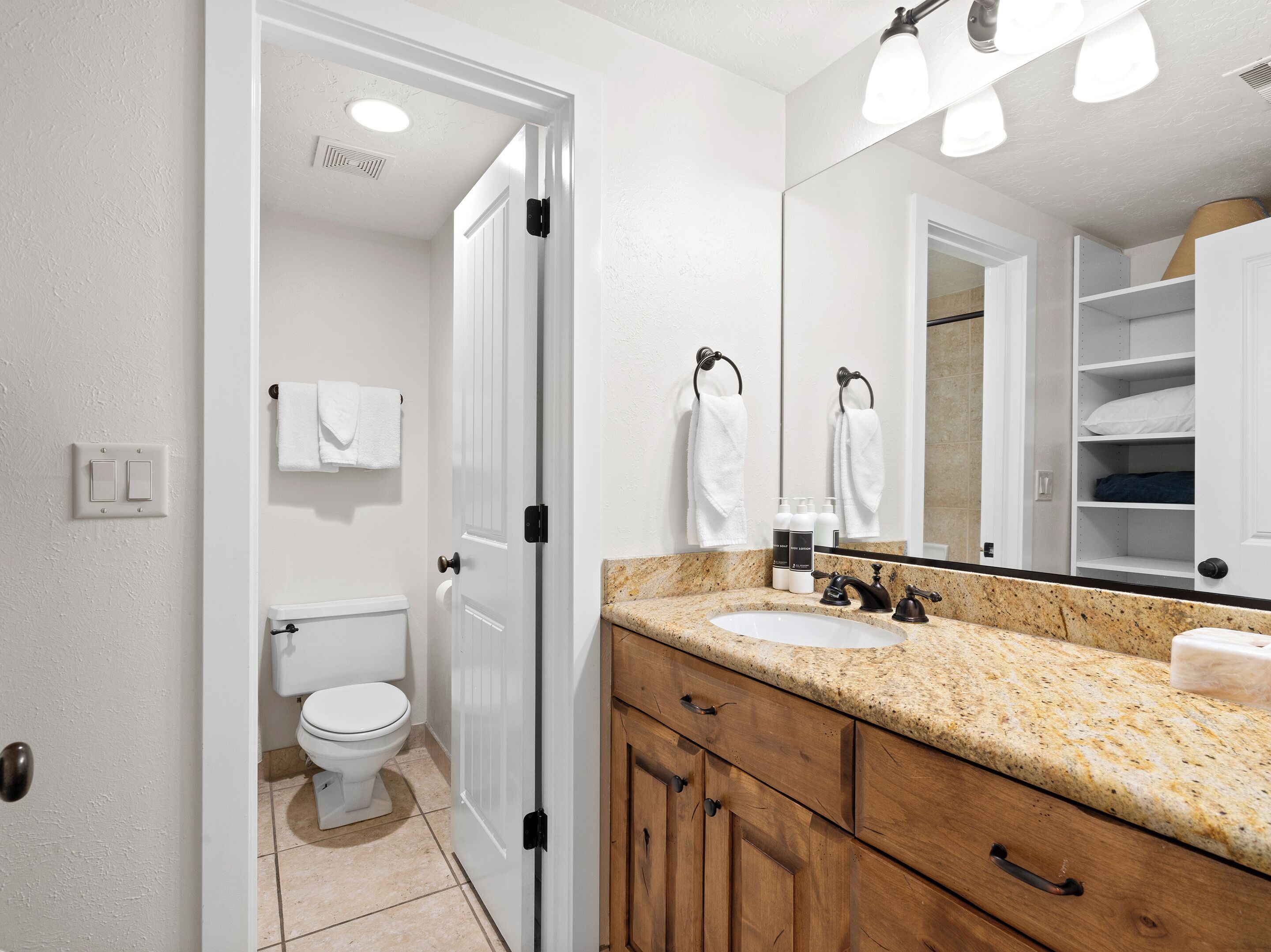 A bathroom with a white toilet, a sink with a granite countertop, wooden cabinets, and a large mirror.