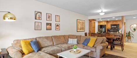A living room with a sectional sofa, a coffee table, and a dining area with wooden cabinets and a kitchen in the background.