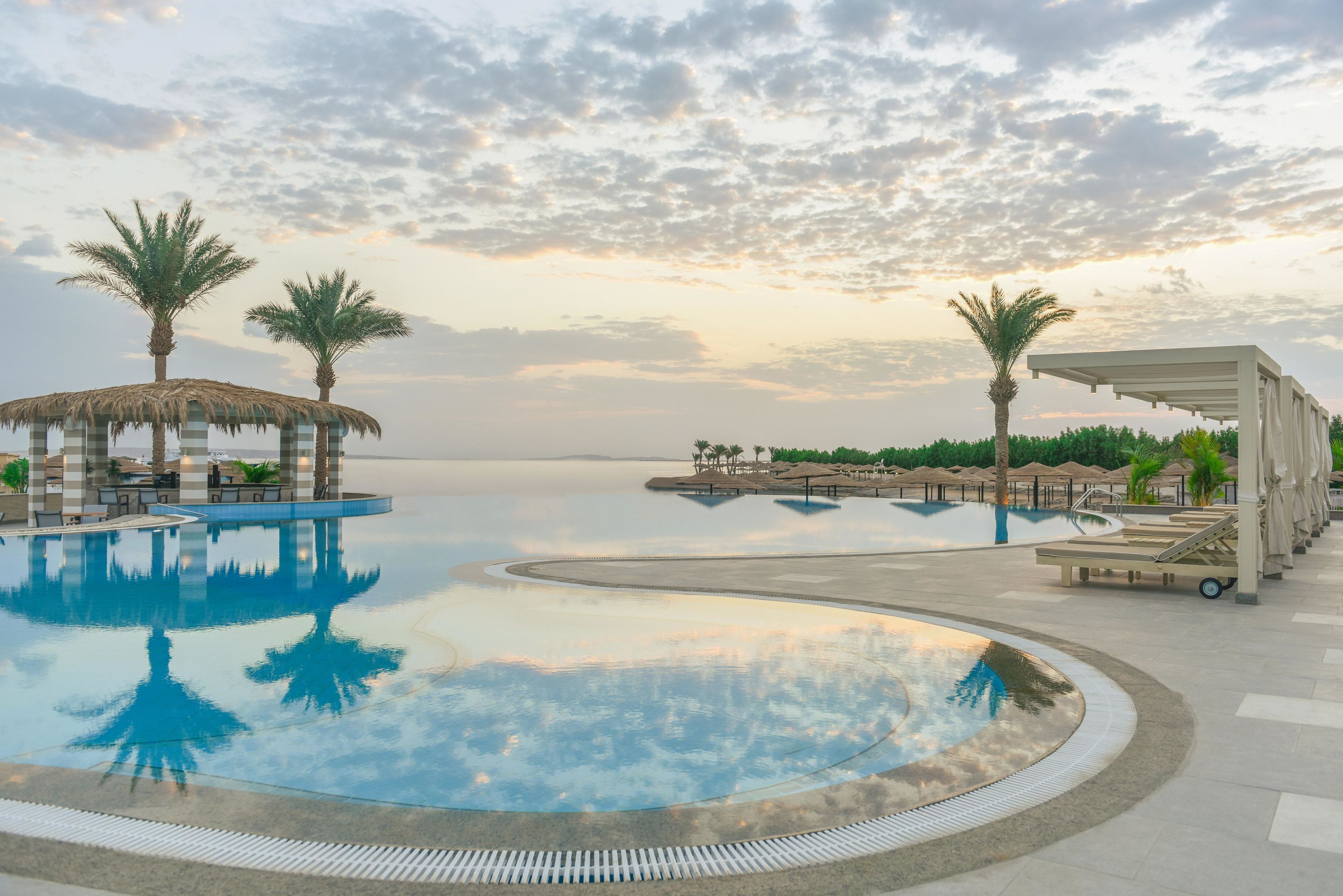A pool with palm trees, lounge chairs, and a covered area.