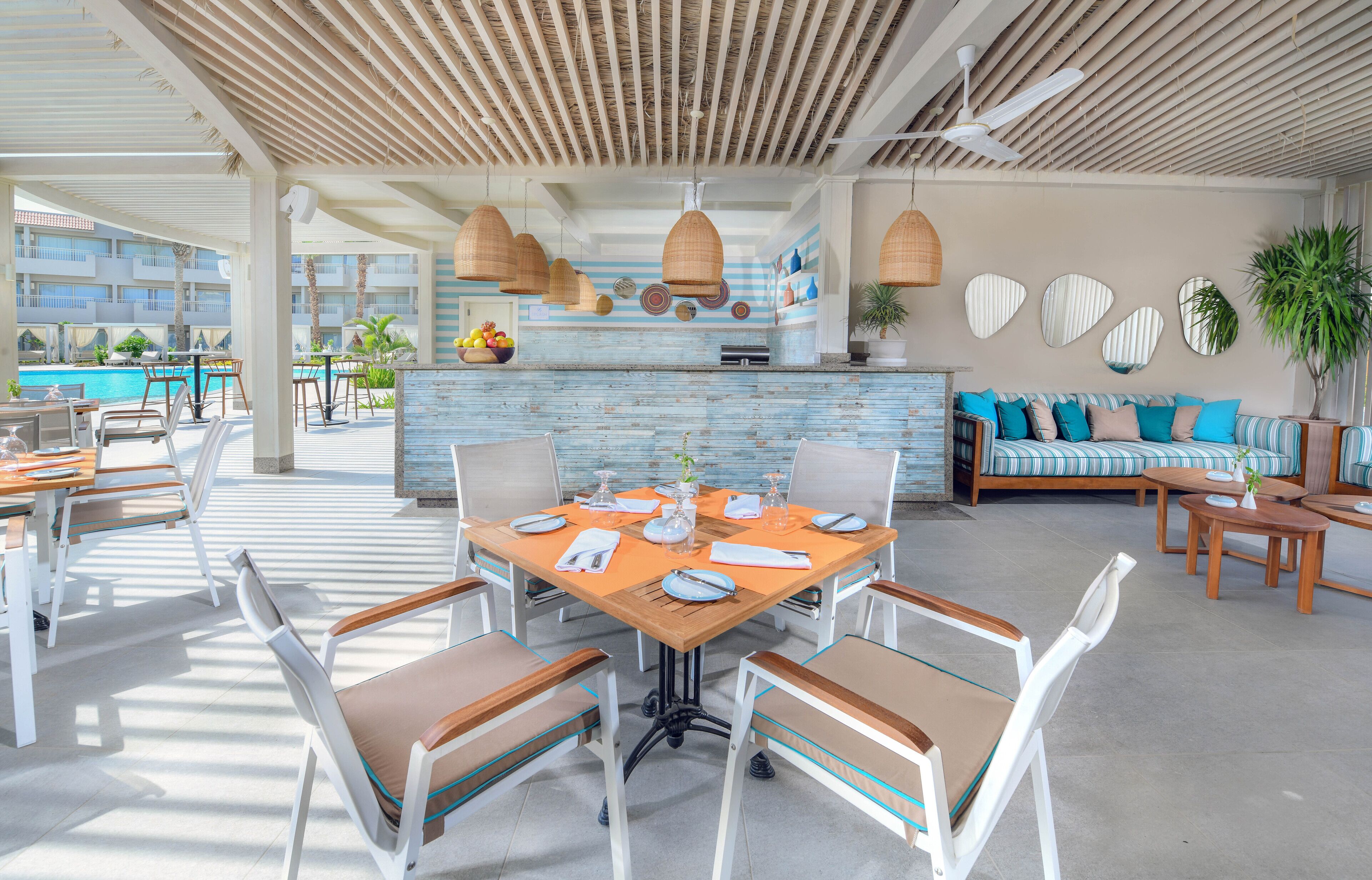 A spacious outdoor dining area with a pool view, a bar with a blue-tiled backsplash, and a seating area with striped cushions.