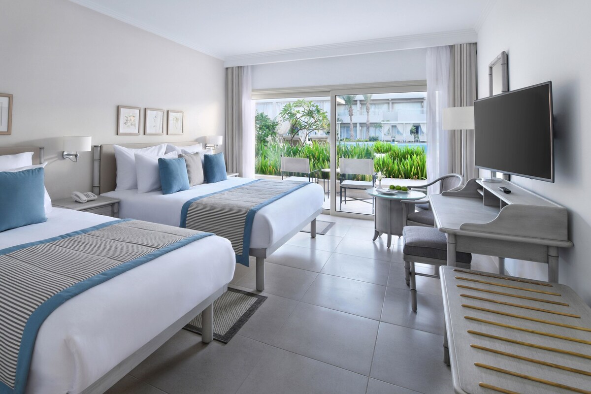 A hotel room with two beds, a desk with a chair, a TV, and a balcony with plants.