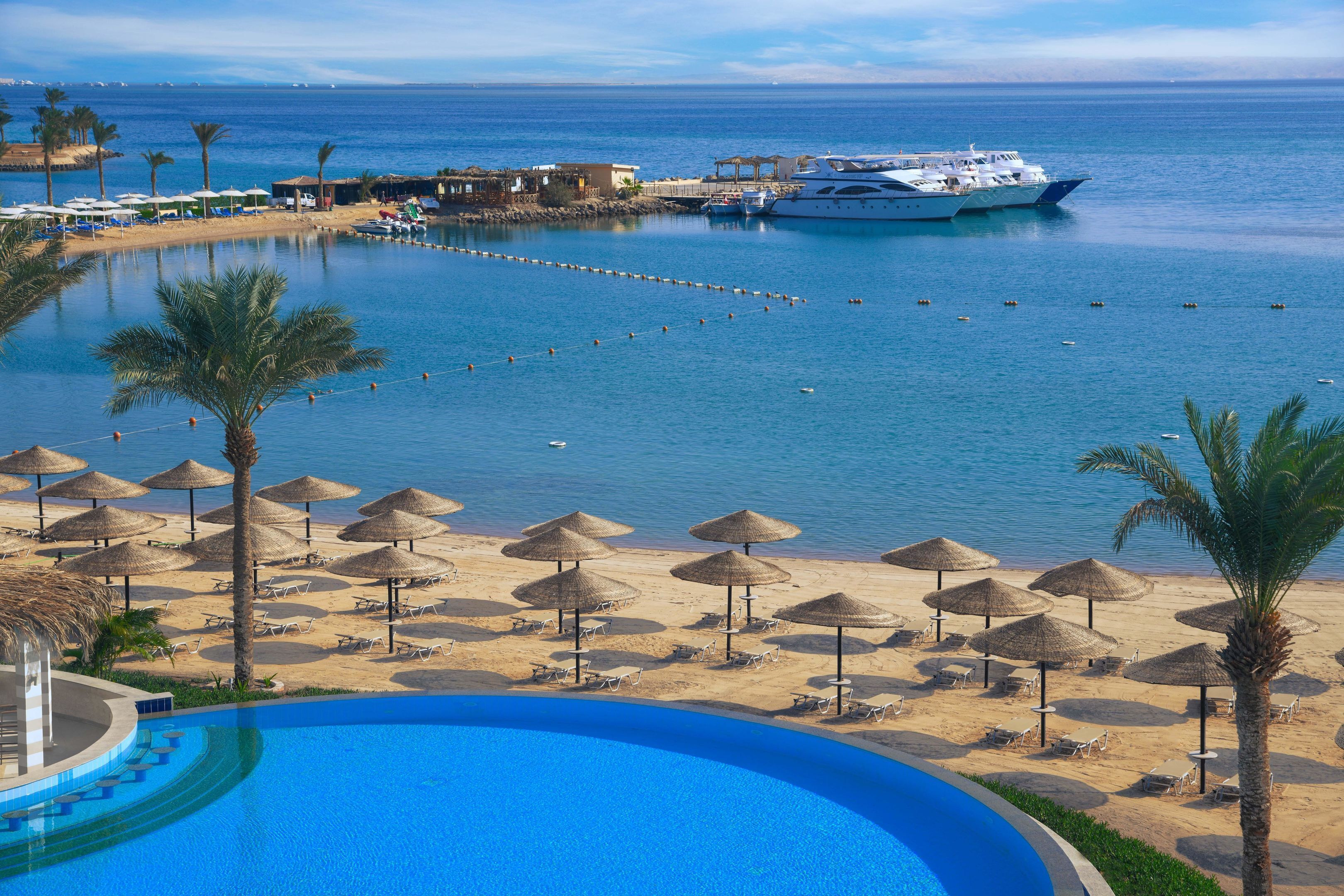 A resort with a swimming pool, beach umbrellas, and a yacht docked near the water.