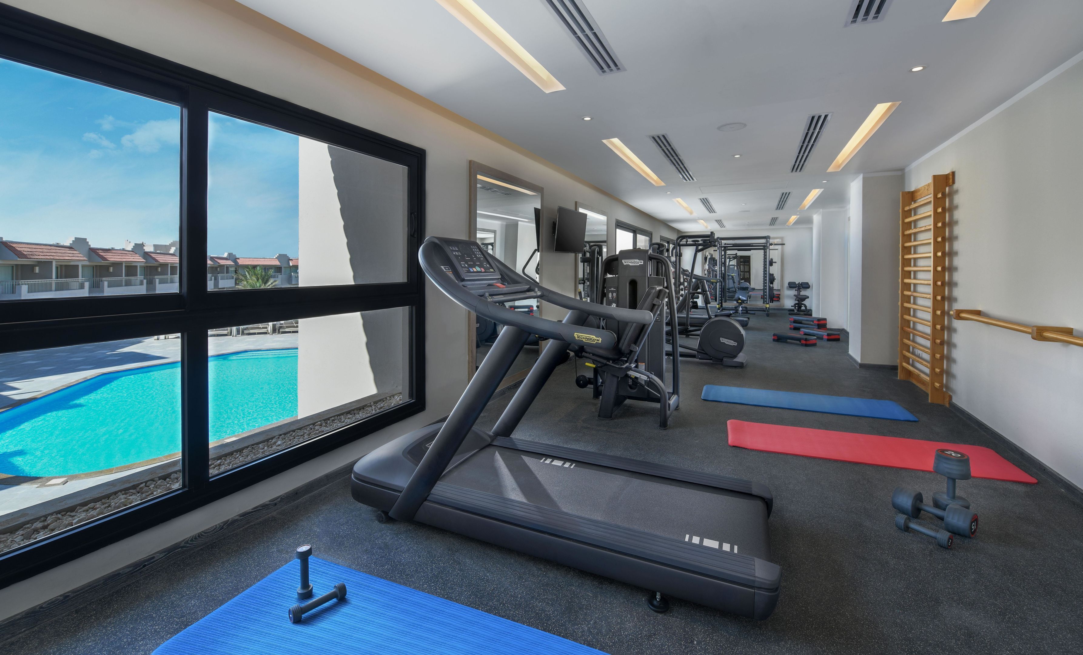 Una palestra con tapis roulant, pesi e materassini da ginnastica, con vista su una piscina.