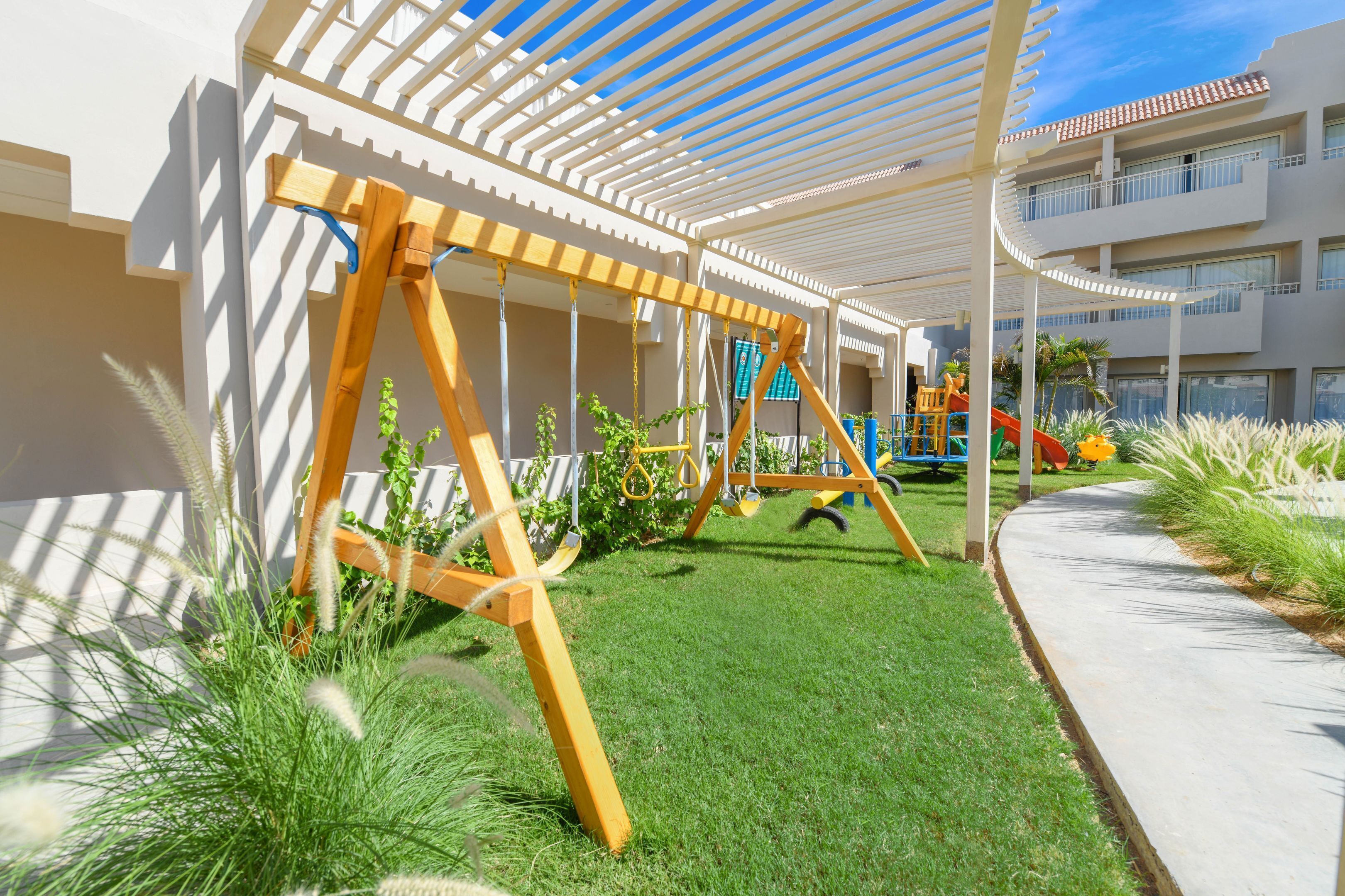 Ein Spielbereich für Kinder mit Schaukeln und Rutsche, umgeben von einer weißen Pergola und einem Rasen.