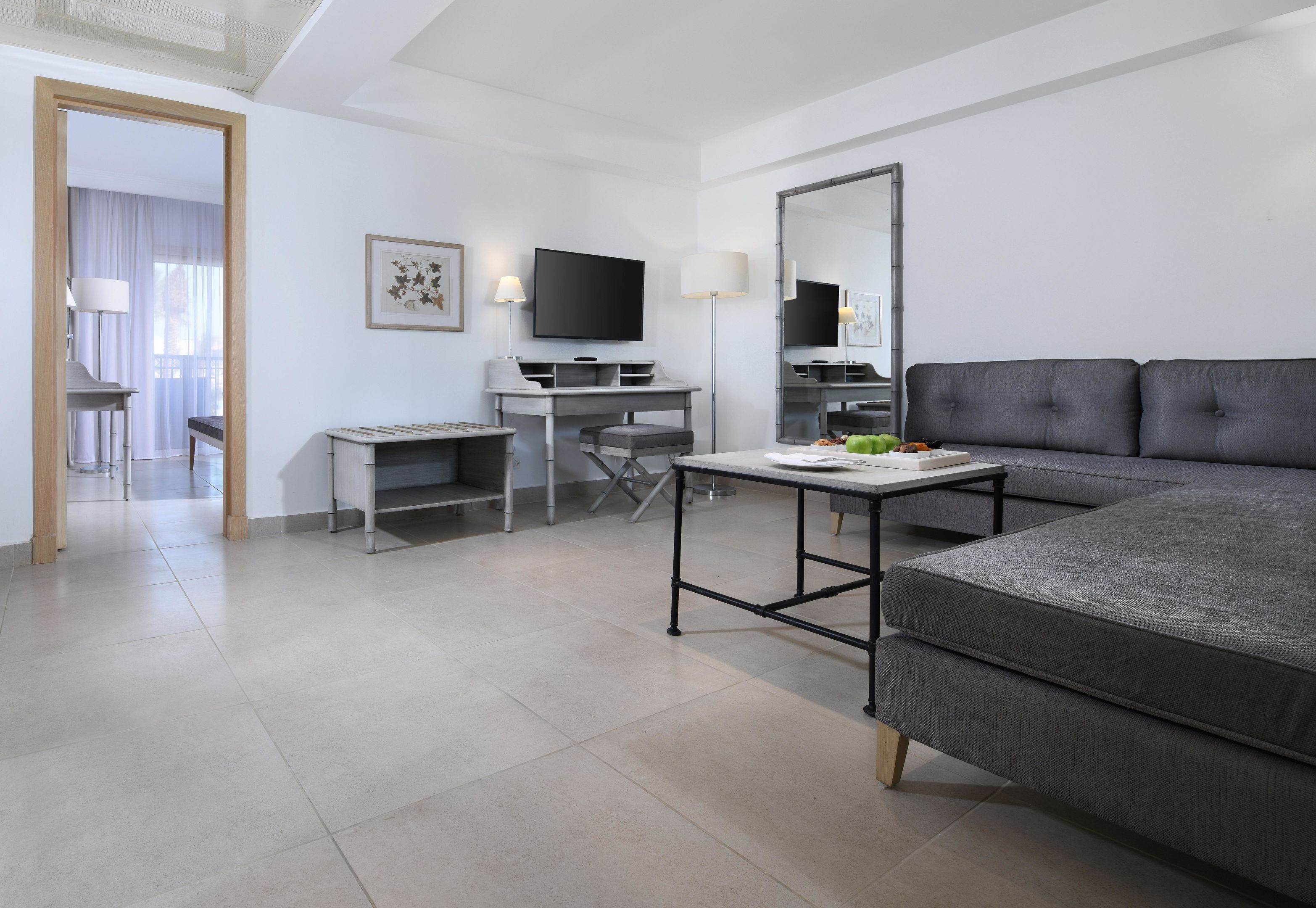 Una habitación de hotel moderna con un sofá gris, un escritorio con televisor de pantalla plana, un espejo grande y una mesa de comedor.