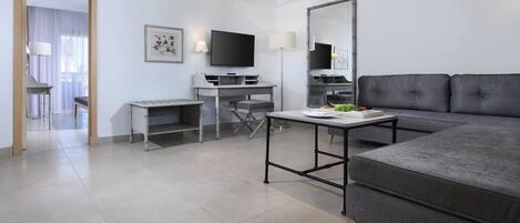 A modern hotel room with a grey sofa, a desk with a flat-screen TV, a large mirror, and a dining table.
