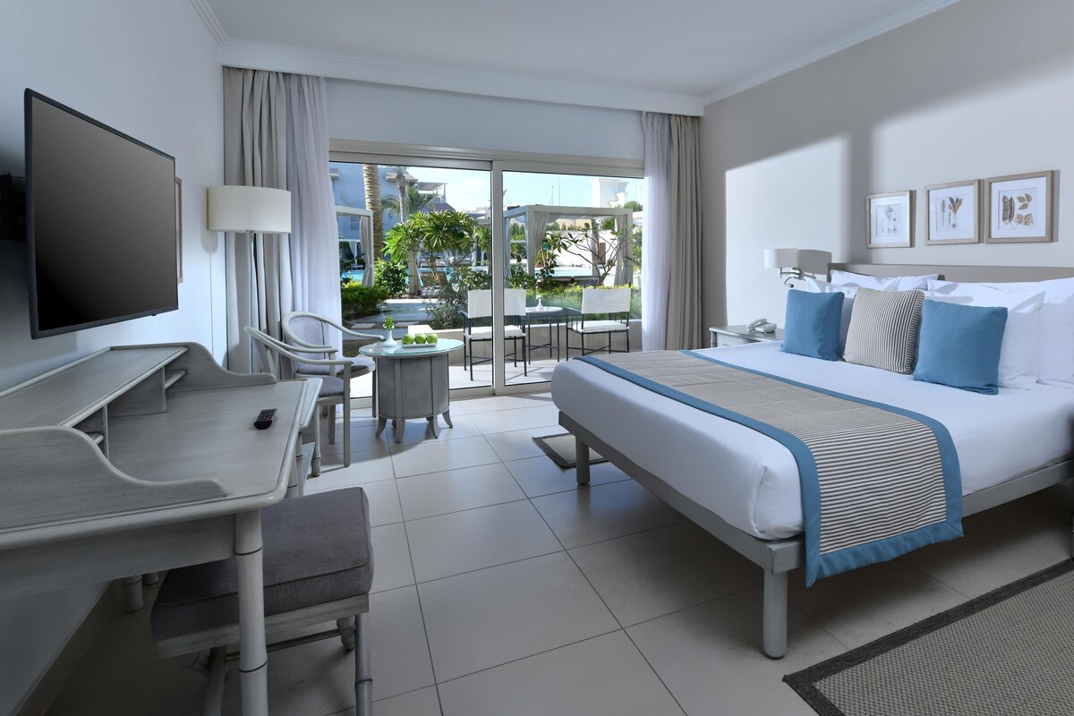 A hotel room with a bed, desk, chair, TV, and a balcony with outdoor seating.