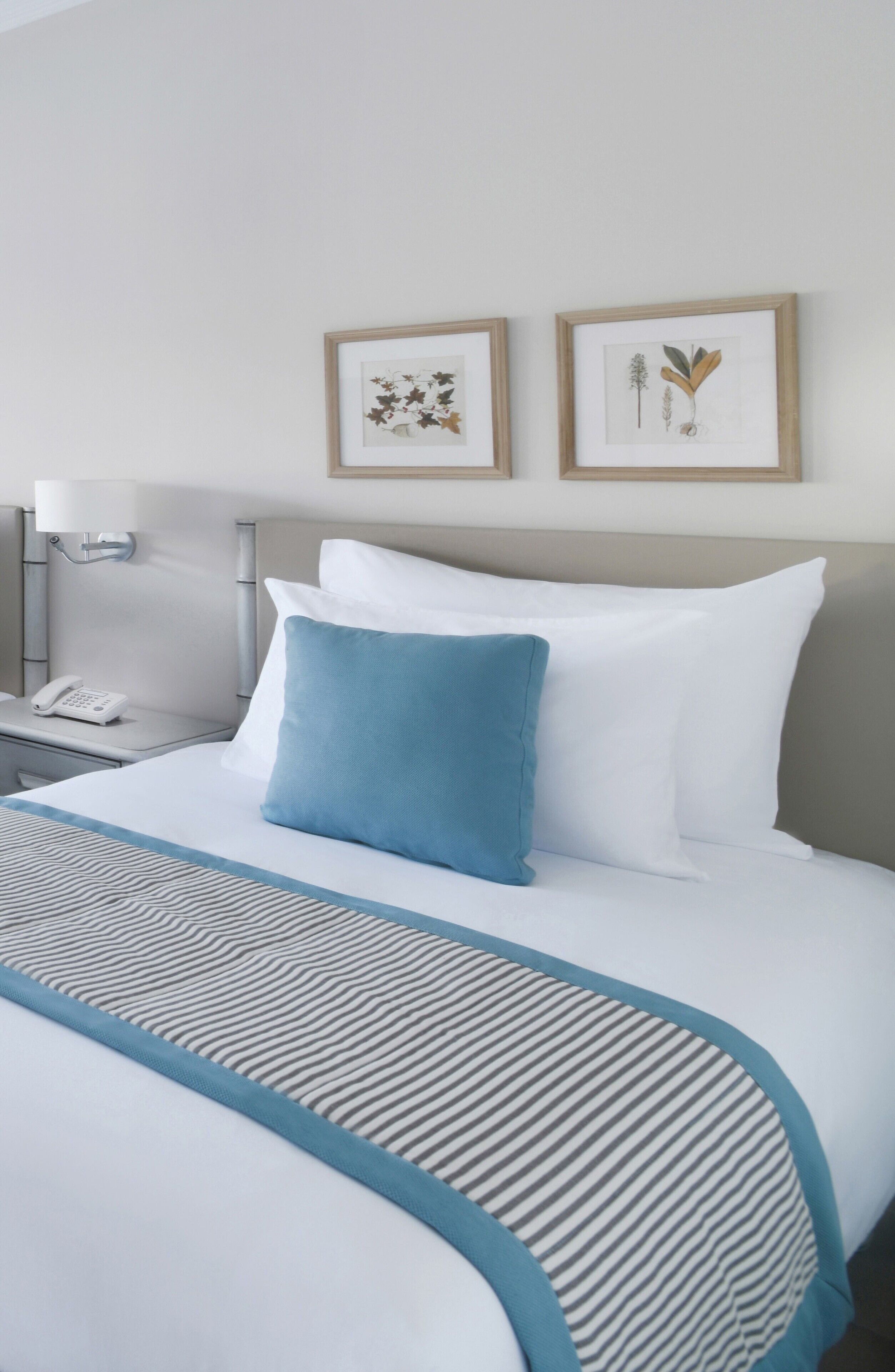 Una habitación de hotel con una cama bien hecha, una almohada decorativa azul y dos láminas botánicas enmarcadas en la pared.