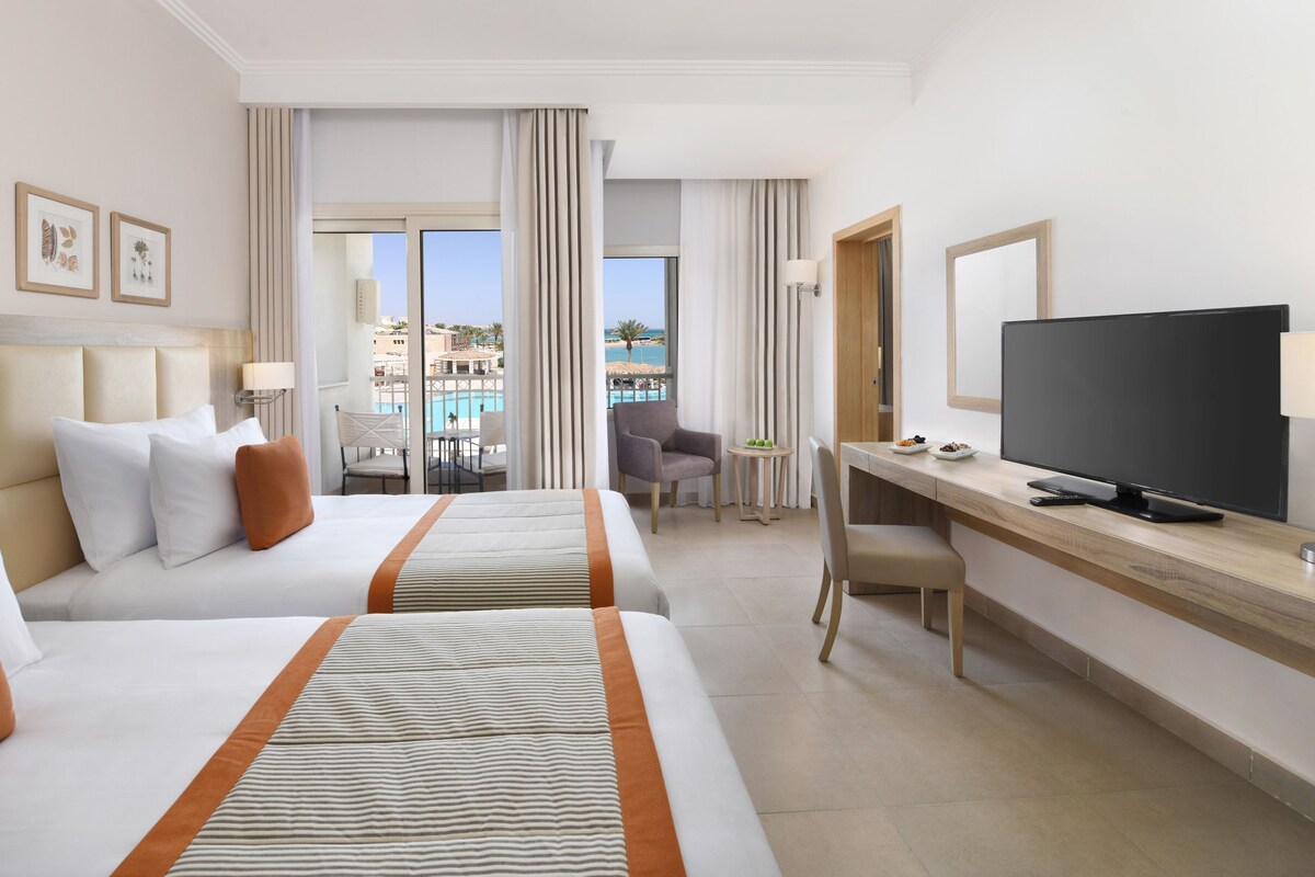 A hotel room with two beds, a desk with a TV, a chair, and a view of a pool and buildings.
