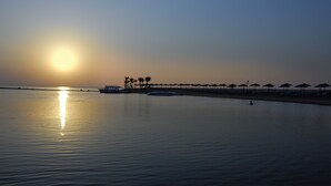 A calm waterfront with a boat docked near the shore, palm trees, and a row of beach umbrellas. - JAZ Elite Casa Del Mar Beach - All inclusive (Hurghada)