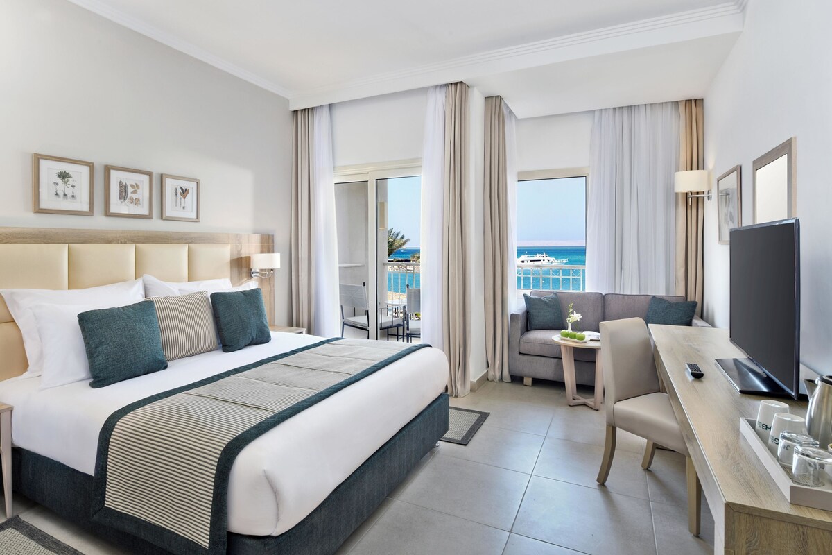 A hotel room with a bed, a desk, a chair, a TV, and a balcony with a view of the sea.