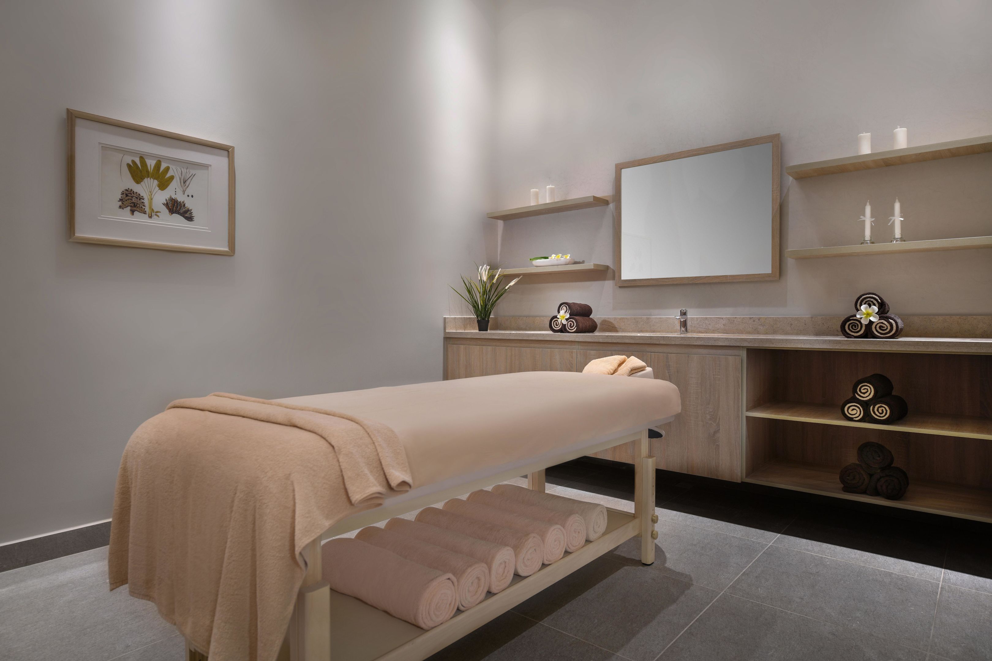 Ein moderner Spa-Raum mit einer Massageliege, Regalen mit Kerzen, einem Spiegel und einem gerahmten Pflanzenprint.