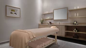 Ein moderner Spa-Raum mit einer Massageliege, Regalen mit Kerzen, einem Spiegel und einem gerahmten Pflanzenprint.