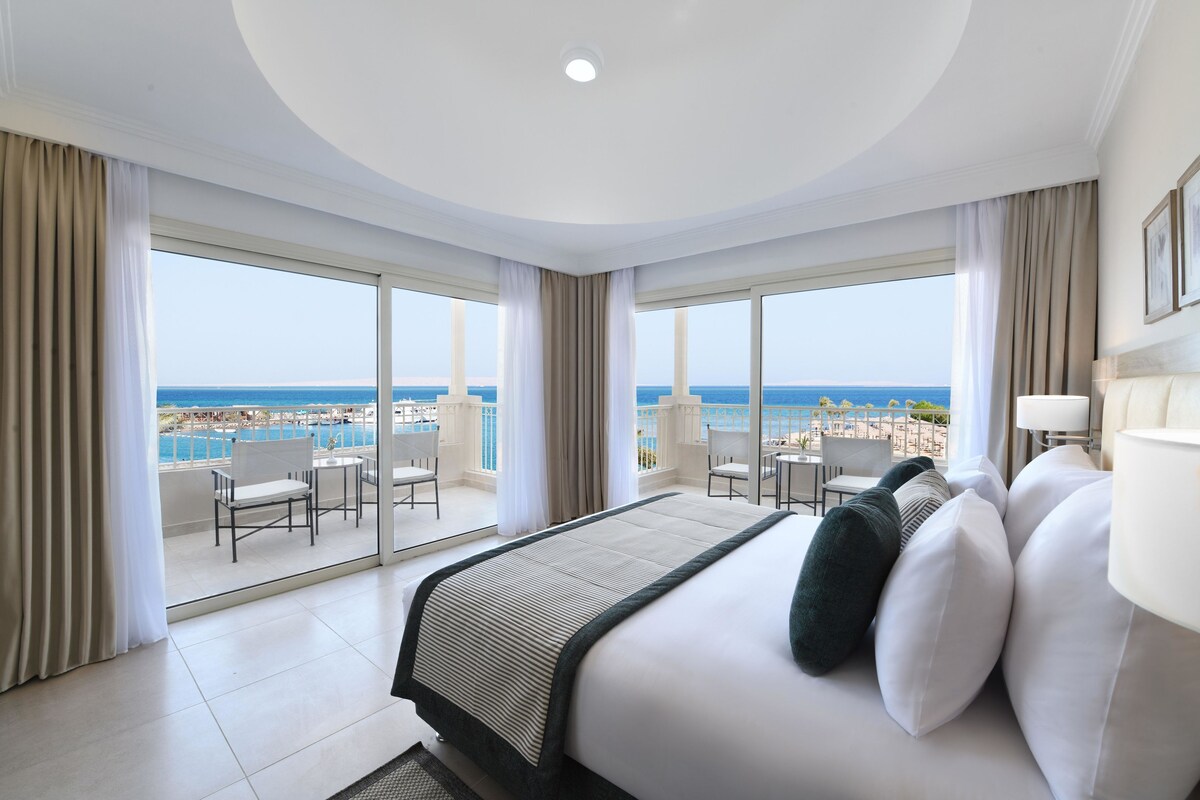 A hotel room with a large bed, a view of the sea, and a balcony with chairs.