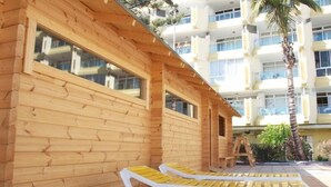 Uma estrutura de madeira com grandes janelas, um deck de madeira com espreguiçadeiras e um edifício alto ao fundo.
