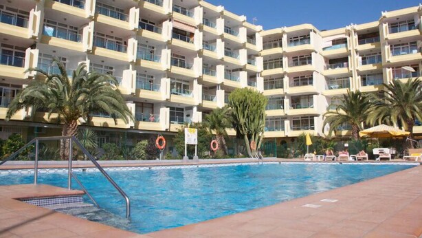 Um hotel com piscina, palmeiras e um edifício de vários andares com varandas.
