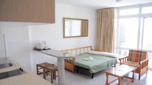 Quarto de hotel compacto com cama, uma pequena mesa, cadeira, cozinha compacta e uma janela grande.