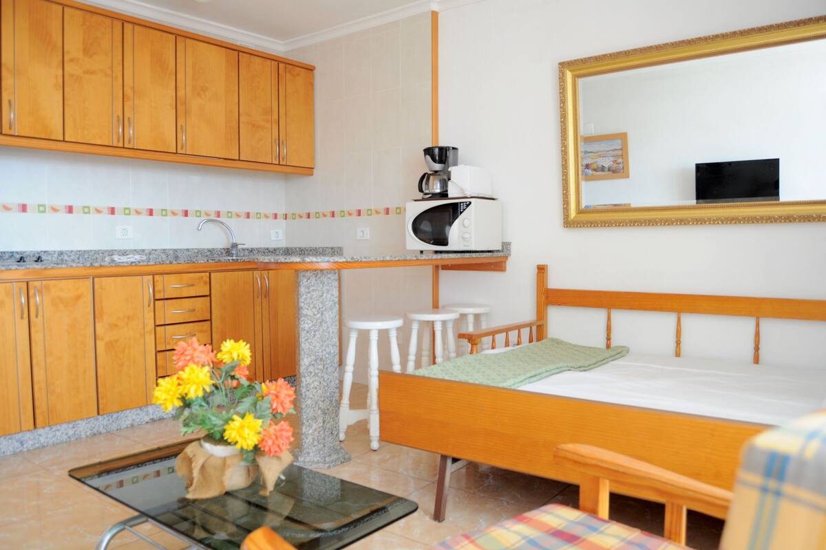 A compact room with a bed, a small kitchen area including a sink and a microwave, and a dining table with stools.